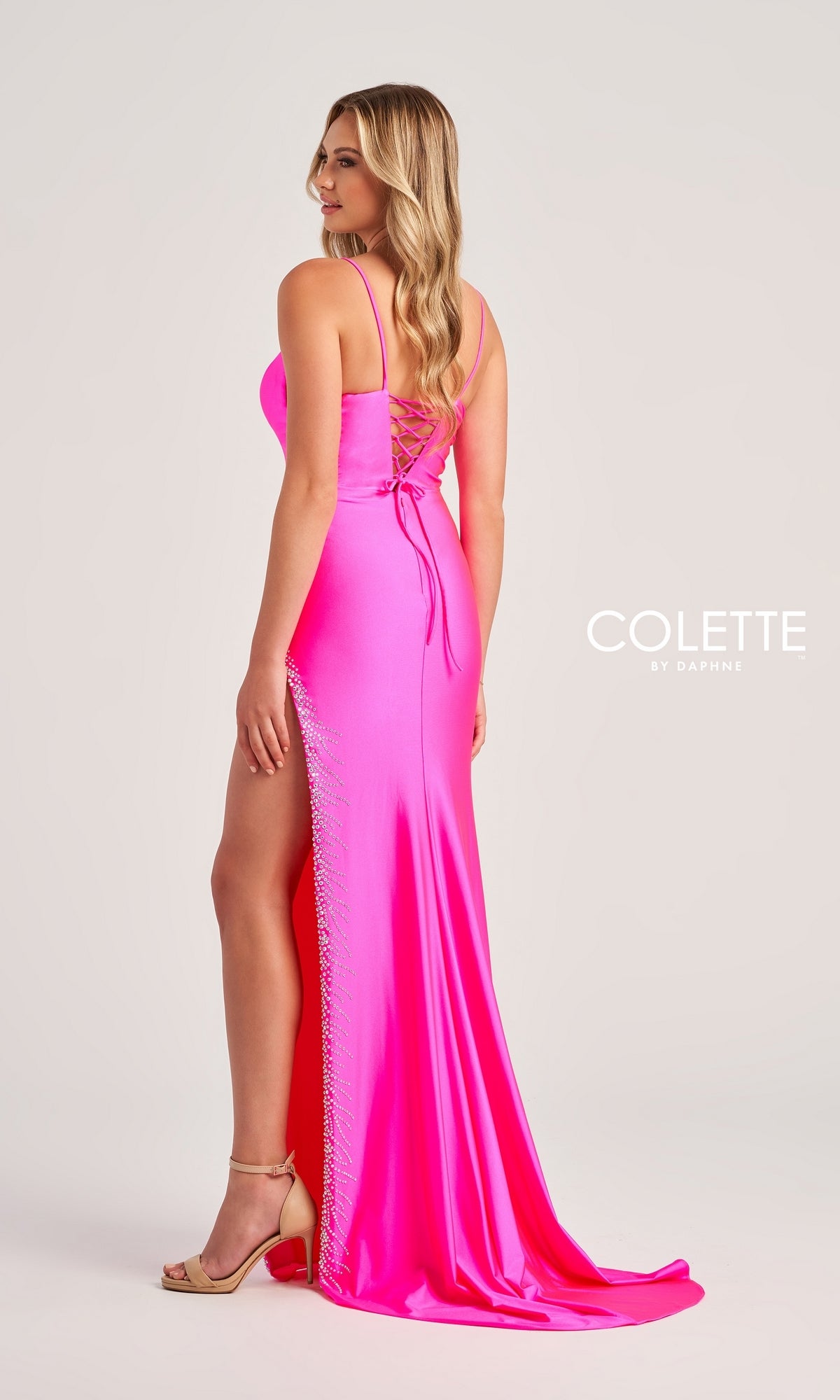 A woman in the Colette Lace-Up Sexy Long Formal Dress CL5200, a bright pink, floor-length gown with a side slit and lace-up back, stands facing away from the camera in nude heels. The